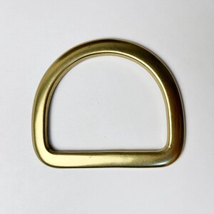 May include: A solid brass D-ring hardware component with a smooth, polished metallic gold finish. The object features a curved top arch and a straight bottom base, forming a classic semi-circular shape used for straps, belts, or bag attachments.