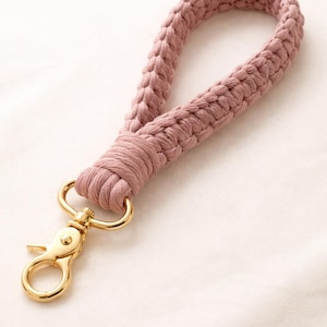 May include: A woven wrist strap in a dusty rose color with a gold-colored clasp. The strap is made of a thick, braided material, and the clasp is a lobster claw style. The strap is designed to be attached to keys or a small bag.