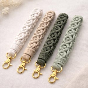 May include: Four macrame wristlet keychains in cream, tan, olive green, and light green. Each keychain features a woven design and a gold-colored lobster clasp for easy attachment to keys or bags.