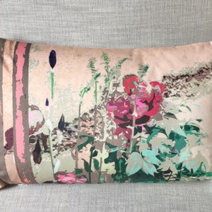 May include: Rectangular cushion featuring a garden scene print. The cushion has a beige background with a colourful illustration of flowers, foliage, and trees in shades of red, green, pink, and brown. The cushion is resting on a grey surface.