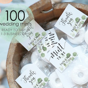May include: A close-up of a wooden bowl filled with small clear bags containing white round mints and personalized wedding favor tags. The tags are decorated with green watercolor leaves and the text "Mint to be" and "Thank you" with the couple's names and wedding date.
