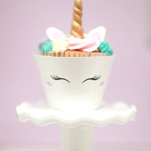 May include: A white cupcake with pink and blue frosting and a gold unicorn horn on top. The cupcake is sitting on a white cake stand.