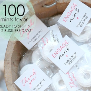 May include: A wooden bowl filled with individually wrapped white mints. Small white tags with pink text that reads "Engage Mint" and "Thank you" are attached. The text "100 mints favor" is also visible.