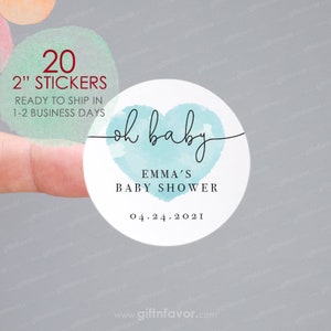May include: Round sticker with the text "oh baby" in elegant script, over a light blue heart design. The sticker also includes the text "EMMA'S BABY SHOWER" and the date "04.24.2021". The sticker is 2 inches in diameter.