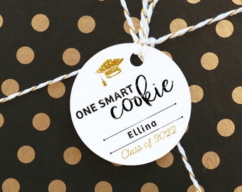 May include: White round gift tag with black text that reads "One Smart Cookie" and a gold glitter graduation cap icon. The tag also has a blank line for a name and the text "Class of 2022".