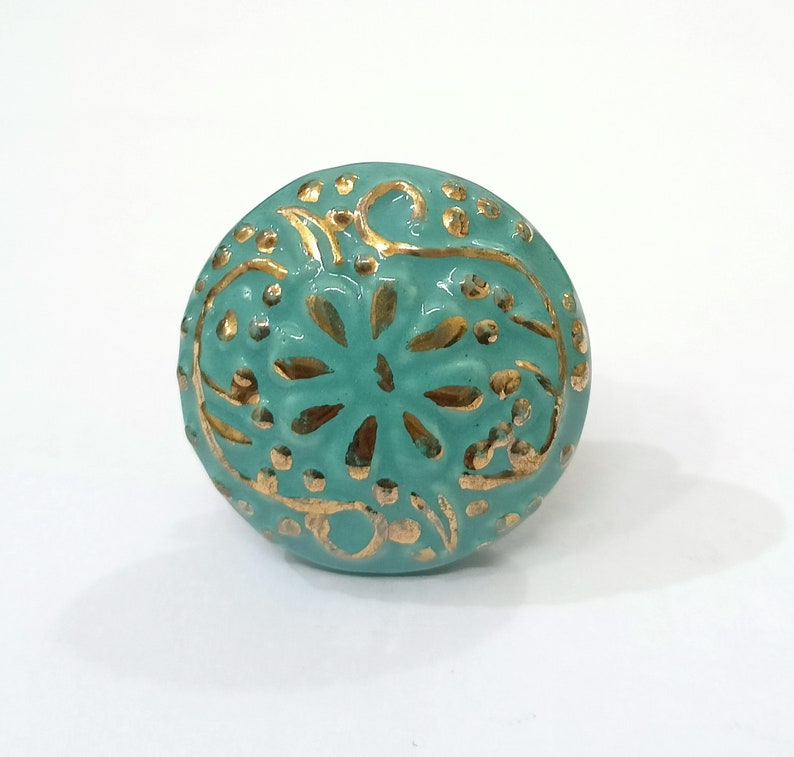 May include: A turquoise blue ceramic knob with a gold floral design. The knob is round and has a raised design.