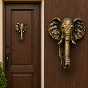May include: A metallic, antique brass-colored door knocker shaped like an elephant head with large ears and a trunk extending downward. The knocker is mounted on a dark brown wooden door with a visible keyhole and a black handle below it.
