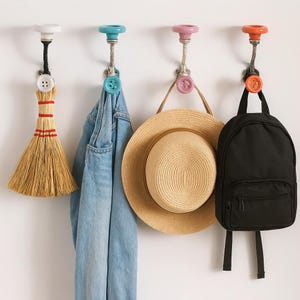 May include: Four decorative wall hooks with colourful knobs and button accents. A small broom, denim jacket, straw hat, and black backpack hang from the hooks. The hooks are mounted on a white wall, offering a stylish storage solution.