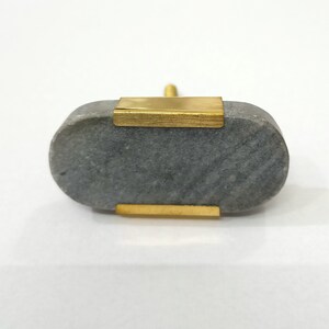 K&ouml;nnte beinhalten: Ein grauer Stein-Schrankknauf mit Messingakzenten. Der ovale Knauf hat Messingplatten auf der Vorder- und R&uuml;ckseite sowie einen Messingstift zur Befestigung. Der Stein hat ein nat&uuml;rliches, strukturiertes Aussehen.