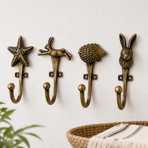Op de afbeelding: Vier antieke messing wandhaken met dierontwerpen: een zeester, een konijn, een egel en een konijn. Elke haak heeft een gebogen onderkant en een decoratieve bovenkant, ideaal om op te hangen.