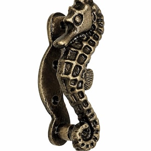 Puede incluir: Un tirador de puerta o mueble con forma de caballito de mar y acabado en bronce oscuro envejecido. El herraje presenta un patr&oacute;n texturizado en cuadr&iacute;cula a lo largo del cuerpo, una cola curvada y una placa de montaje con tres orificios para tornillos.