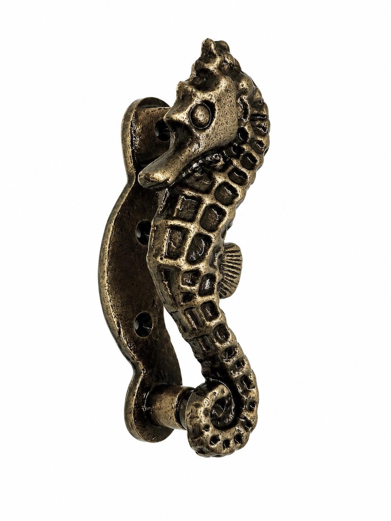 Puede incluir: Un tirador de puerta o mueble con forma de caballito de mar y acabado en bronce oscuro envejecido. El herraje presenta un patr&oacute;n texturizado en cuadr&iacute;cula a lo largo del cuerpo, una cola curvada y una placa de montaje con tres orificios para tornillos.