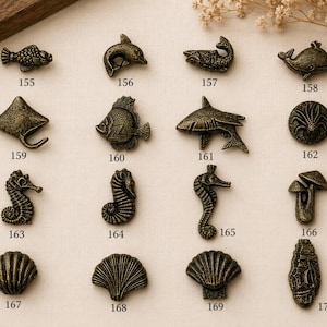 May include: A collection of sixteen bronze-toned metal cabinet knobs shaped like marine life and nature elements, labelled with numbers 155 through 170. The designs include various fish, a dolphin, a whale, a stingray, seahorses, shells, and mushrooms.