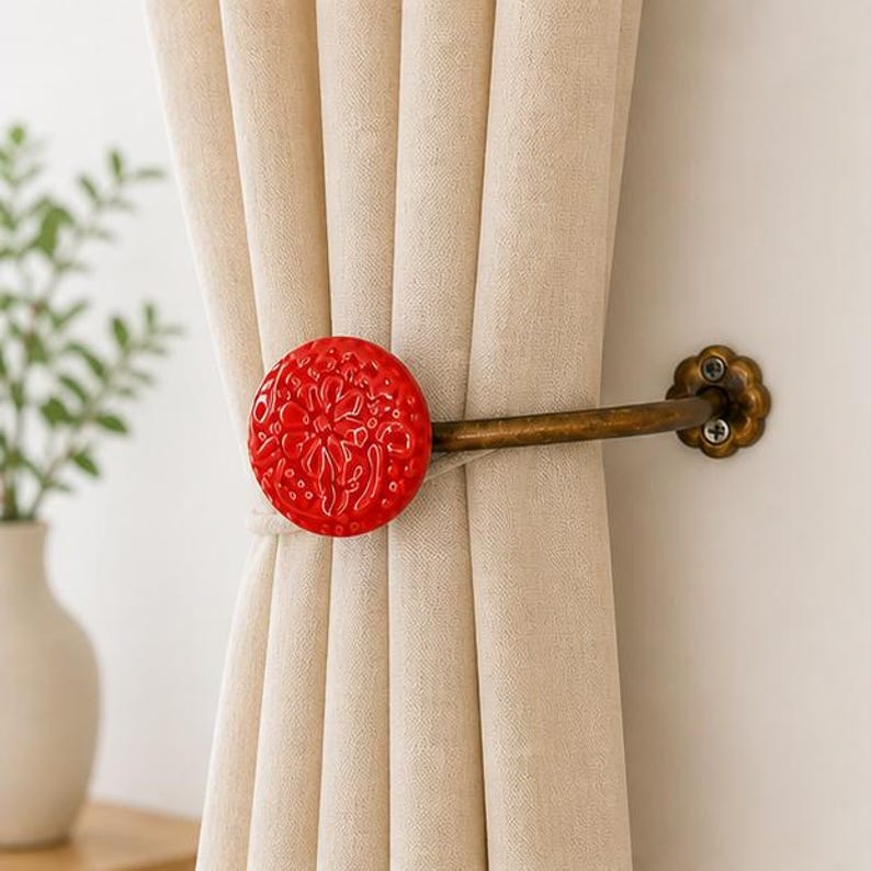 May include: A curtain holdback featuring a bright red circular ceramic knob with an embossed floral pattern, attached to a curved antique brass metal arm that mounts to the wall with a flower-shaped base plate, securing a beige fabric curtain.