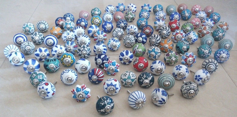 Op de afbeelding: Een verzameling van 50 keramische kastknoppen met verschillende kleurrijke bloemmotieven. De knoppen zijn wit met blauwe, groene, rode en gele accenten. Elke knop heeft een zilveren metalen basis.