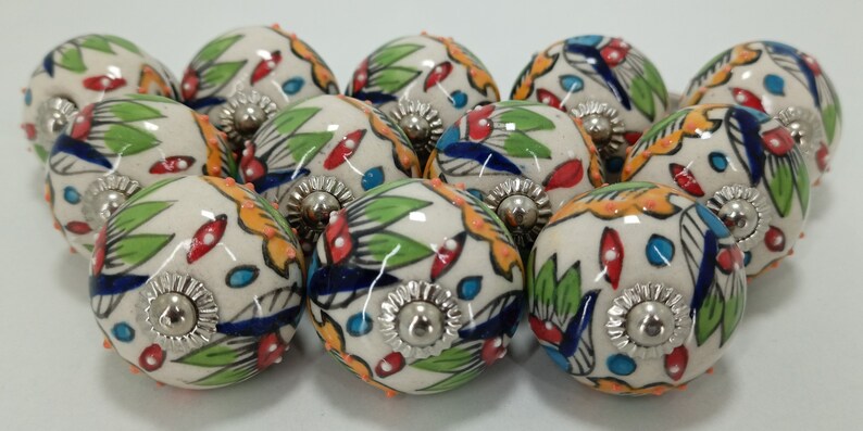 K&ouml;nnte beinhalten: Eine Sammlung von 12 Keramik-Schubladengriffen mit wei&szlig;er Basis und bunten Blumenmustern. Die Griffe haben einen silbernen Metallakzent in der Mitte.