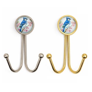 Puede incluir: Dos perchas decorativas de pared. Una es plateada, la otra dorada. Cada percha presenta un diseño circular con un pájaro azul posado en una rama florecida. Las perchas tienen un diseño de doble gancho con una bola redondeada al final de cada gancho.