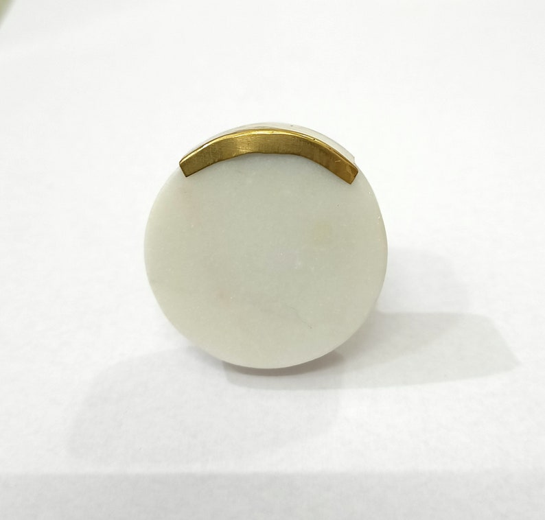 K&ouml;nnte beinhalten: Runder, wei&szlig;er Marmor-Schrankknopf mit einem gebogenen, goldfarbenen Metallakzent. Der Knopf ist glatt und poliert, mit einem einfachen, eleganten Design. Diese Hardware ist f&uuml;r Schubladen und Schr&auml;nke geeignet.