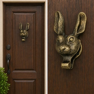 May include: A brass-toned metal door knocker shaped like a rabbit head with long, upright ears and a textured finish. The rabbit head is mounted on a dark brown wooden door, featuring a hinged lower section that serves as the striking mechanism.