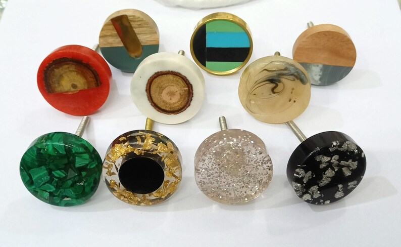 May include: Assortment of 10 decorative round cabinet knobs with various designs and colors. The knobs feature a variety of materials, including wood, resin, and metal. Some knobs have a glittery finish, while others have a smooth, polished surface.