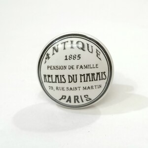 May include: White ceramic knob with a black circle around the edge. The circle has the text "Antique 1885 Pension de Famille Relais du Marais 73, Rue Saint Martin Paris" in black.