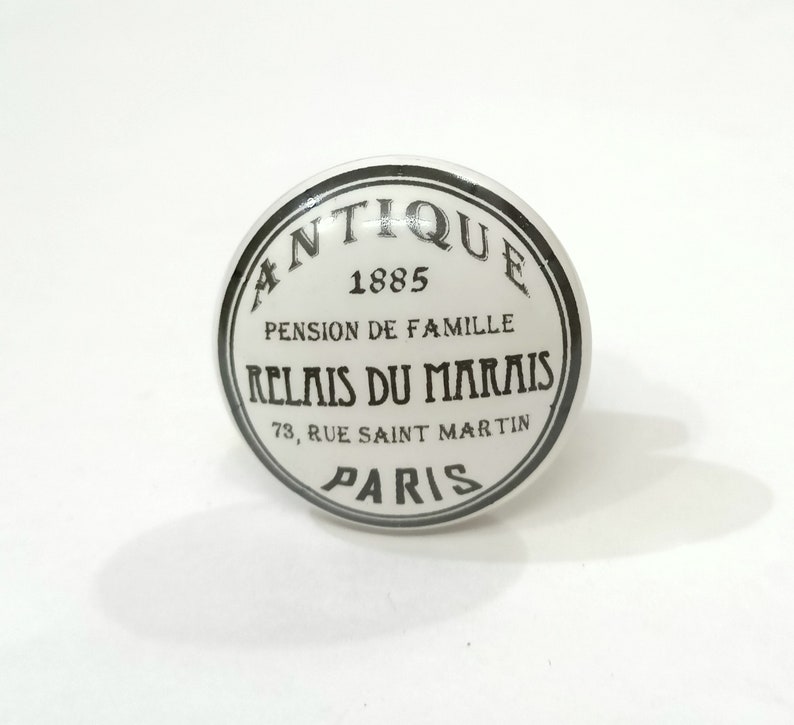 May include: White ceramic knob with a black circle around the edge. The circle has the text "Antique 1885 Pension de Famille Relais du Marais 73, Rue Saint Martin Paris" in black.