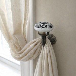 May include: A decorative curtain hook with a white ceramic knob featuring a black floral design. The hook is mounted on a wall and holds a cream-coloured curtain. The hook has a bronze finish.