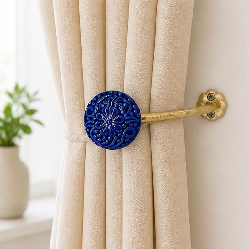 May include: A curtain tieback featuring a circular, deep blue ceramic disc with an intricate embossed floral pattern. The disc is attached to a curved, metallic gold-toned arm that mounts to the wall with a flower-shaped base plate.