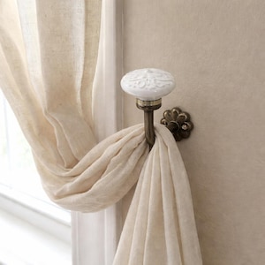 May include: A decorative curtain tieback hook with a white ceramic knob and a bronze-coloured metal base. The hook is holding a cream-coloured curtain. The knob has a detailed floral design.