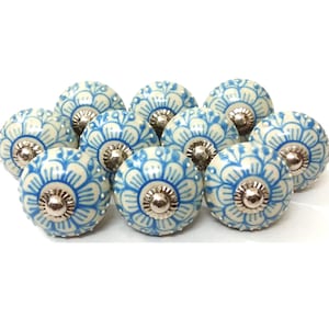 Puede incluir: Un conjunto de pomos de cerámica para armarios. Cada pomo presenta una base crema con un diseño floral azul y un centro de metal plateado. Los pomos son redondos y tienen una estética decorativa y vintage.