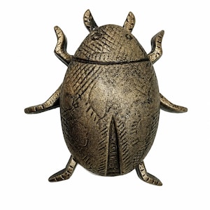Puede incluir: Una figura de escarabajo en tono bronce con una superficie texturizada y desgastada. El objeto con forma de insecto presenta patrones lineales grabados en su espalda redondeada, dos antenas en la parte superior y seis patas extendidas.