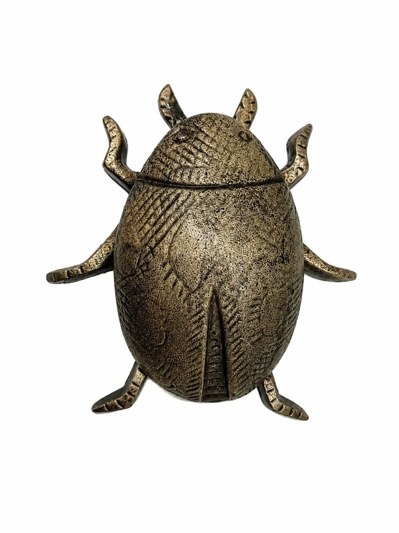 Puede incluir: Una figura de escarabajo en tono bronce con una superficie texturizada y desgastada. El objeto con forma de insecto presenta patrones lineales grabados en su espalda redondeada, dos antenas en la parte superior y seis patas extendidas.