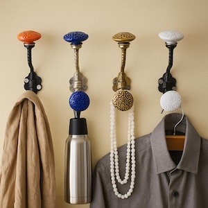 May include: Four decorative wall hooks with ornate ceramic knobs in orange, blue, gold, and white. Each hook has a black or silver metal base. A tan coat, a water bottle, a necklace, and a gray shirt hang from the hooks.