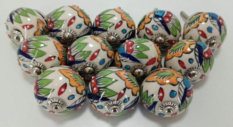 K&ouml;nnte beinhalten: Zw&ouml;lf Keramik-Kn&ouml;pfe mit wei&szlig;em Hintergrund und bunten Blumenmustern. Die Kn&ouml;pfe haben eine silberne Metallbasis.