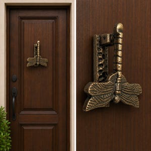 May include: A bronze-toned metal door knocker shaped like a dragonfly with textured wings and a segmented body. The knocker is mounted on a dark brown wooden door featuring a vertical panel design, a black handle, and a matching black keyhole plate.
