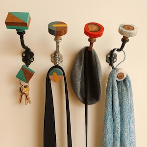 Puede incluir: Cuatro percheros de pared decorativos con diseños únicos. Los ganchos presentan tapas de madera en varias formas y colores, incluyendo turquesa, rojo y gris. Un gancho sostiene llaves, mientras que otros exhiben una corbata negra, una bufanda gris y una bufanda azul paisley.
