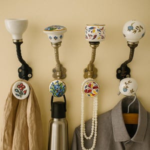 May include: Four decorative wall hooks with ceramic knobs and metal stems. The knobs feature floral and abstract designs in various colors. One hook holds a tan scarf, another a water bottle, a third a pearl necklace, and the last a gray jacket.