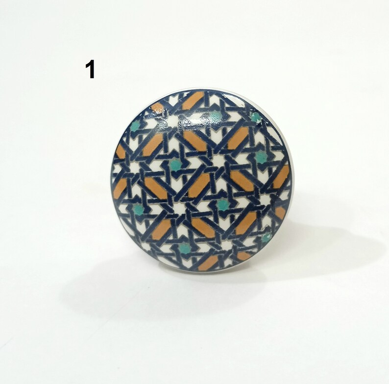 Może przedstawiać: Okrągła ceramiczna gałka do szafki z geometrycznym wzorem. Wz&oacute;r składa się z zazębiających się kształt&oacute;w w kolorze granatowym, pomarańczowym, turkusowym i białym. Numer "1" jest wydrukowany na g&oacute;rze po lewej stronie gałki.