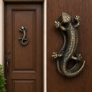 May include: A bronze-colored metal door knocker shaped like a lizard with a textured, scaly surface. The lizard is depicted in a climbing position with its head facing upward and its tail curved downward, mounted on a dark brown wooden door.