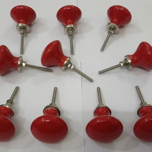 K&ouml;nnte beinhalten: Zehn rote Keramik-Schrankkn&ouml;pfe mit silbernen Metallschrauben. Die Kn&ouml;pfe haben eine abgerundete, leicht gew&ouml;lbte Form.