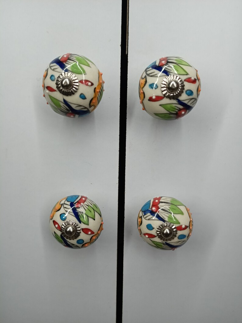 K&ouml;nnte beinhalten: Vier Keramik-Schrankkn&ouml;pfe mit wei&szlig;em Hintergrund und bunten Blumenmustern. Die Kn&ouml;pfe haben eine silberne Metallbasis.