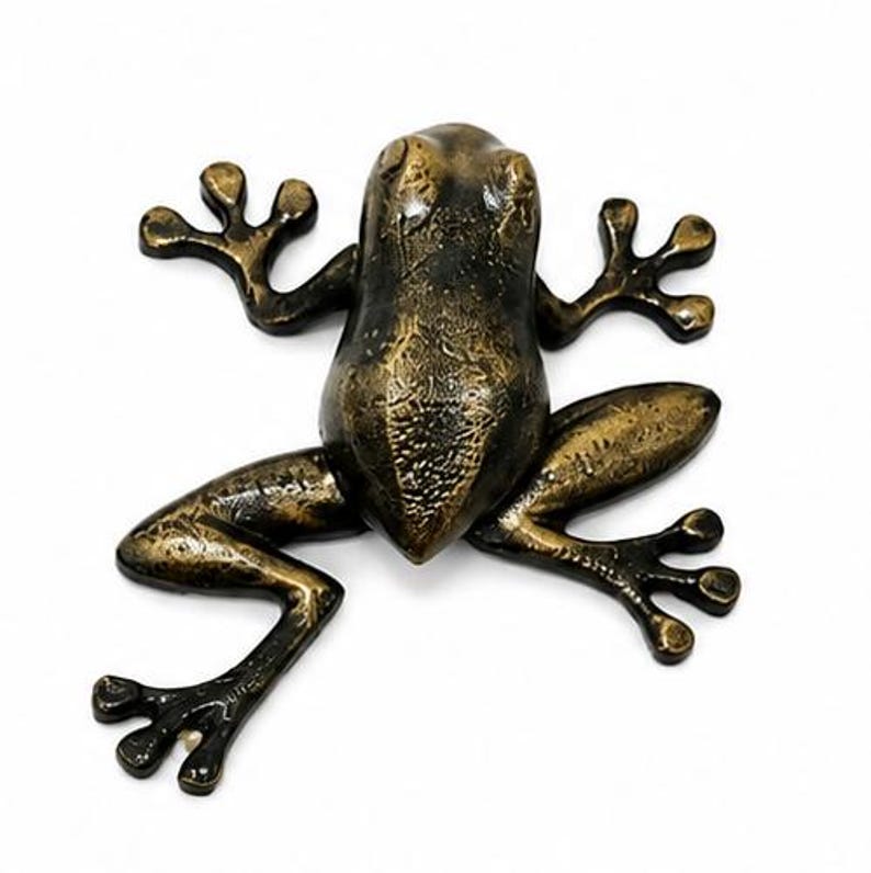 Puede incluir: Una figura de rana de metal color bronce con un acabado texturizado y desgastado. La rana se muestra en una posici&oacute;n plana y extendida con cuatro patas abiertas, dedos visibles y un dorso liso y redondeado con una p&aacute;tina oscura y dorada.