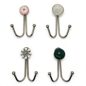 Puede incluir: Cuatro colgadores de pared decorativos con marcos de metal plateado y ganchos dobles. Cada colgador presenta una parte superior de cerámica diferente: un diseño rojo y blanco, un diseño blanco y dorado, un diseño de flor blanca y una esfera verde oscuro.