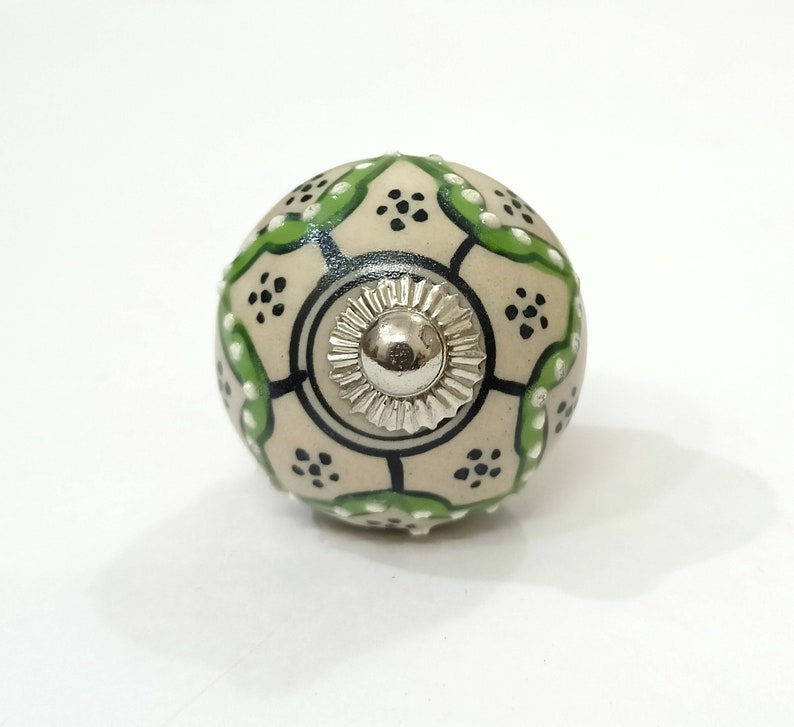 Op de afbeelding: Een witte keramische knop met een geometrisch patroon in groen en zwart. De knop heeft een zilveren metalen midden.