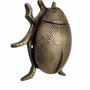 Puede incluir: Una figura de escarabajo de metal en tono bronce con un patr&oacute;n de caparaz&oacute;n texturizado de l&iacute;neas cruzadas y patas articuladas. El objeto con forma de insecto tiene un acabado met&aacute;lico desgastado con p&aacute;tina oscura visible en las hendiduras.