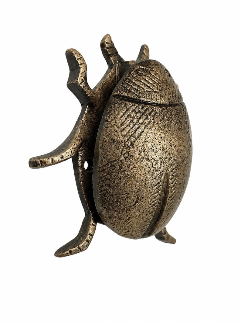 Puede incluir: Una figura de escarabajo de metal en tono bronce con un patr&oacute;n de caparaz&oacute;n texturizado de l&iacute;neas cruzadas y patas articuladas. El objeto con forma de insecto tiene un acabado met&aacute;lico desgastado con p&aacute;tina oscura visible en las hendiduras.