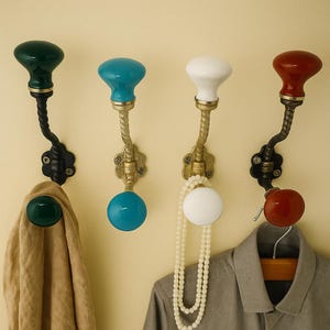 Könnte beinhalten: Vier dekorative Wandhaken mit farbigen Keramikknöpfen. Die Haken haben ein gedrehtes Metalldesign und sind an einer hellen Wand montiert. Ein Haken hält einen beigen Mantel, ein weiterer eine Perlenkette und ein weiteres ein graues Hemd.