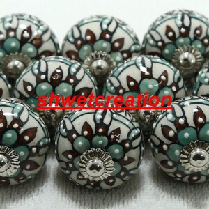 Op de afbeelding: Een set van tien keramische kastknoppen met een witte achtergrond en een bloemmotief in tinten bruin en teal. De knoppen hebben een zilveren metalen basis.