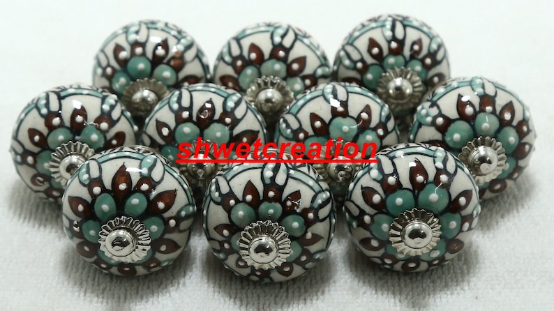 Op de afbeelding: Een set van tien keramische kastknoppen met een witte achtergrond en een bloemmotief in tinten bruin en teal. De knoppen hebben een zilveren metalen basis.