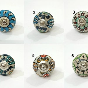 Op de afbeelding: Zes keramische knoppen met verschillende bloemenpatronen. De knoppen zijn wit met blauwe, groene, oranje en rode accenten. De knoppen hebben een diameter van ongeveer 2,5 cm.